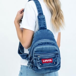 K&ouml;nnte beinhalten: Blauer Jeans-Rucksack mit verstellbaren Tr&auml;gern und einer Vordertasche. Der Rucksack hat das rote Levi's-Logo-Patch. Die Tasche hat eine abgerundete Form und einen Quasten-Rei&szlig;verschluss.