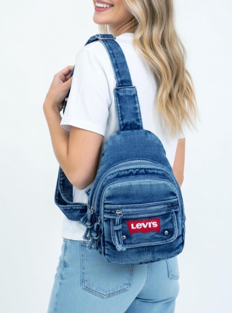 K&ouml;nnte beinhalten: Blauer Jeans-Rucksack mit verstellbaren Tr&auml;gern und einer Vordertasche. Der Rucksack hat das rote Levi's-Logo-Patch. Die Tasche hat eine abgerundete Form und einen Quasten-Rei&szlig;verschluss.