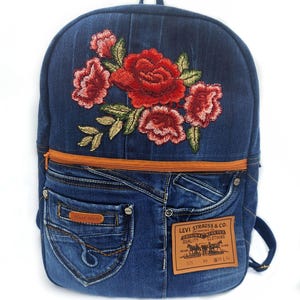 Puede incluir: Una mochila de mezclilla azul oscuro con un gran bordado floral rojo en la parte superior. El frente incluye una cremallera naranja horizontal, un bolsillo pequeño con una etiqueta marrón que dice Hand Made y un parche de cuero beige.