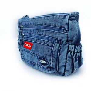 May include: A blue denim crossbody bag featuring multiple zipped compartments and a red rectangular patch with the white text Levi's. The bag includes side pockets with elasticated openings and a small silver-toned metallic accent on the front.