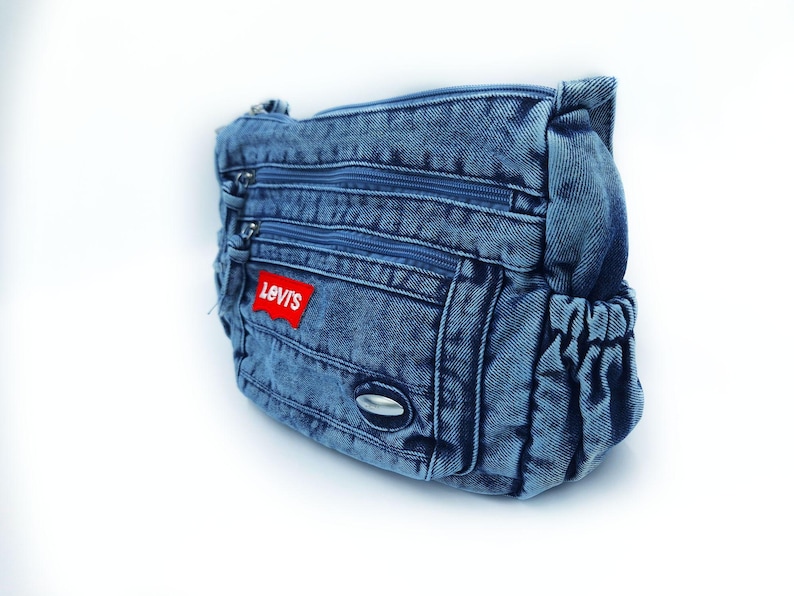 May include: A blue denim crossbody bag featuring multiple zipped compartments and a red rectangular patch with the white text Levi's. The bag includes side pockets with elasticated openings and a small silver-toned metallic accent on the front.