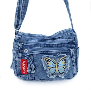 May include: A blue denim crossbody bag featuring multiple zipped compartments, a long adjustable shoulder strap, and a red Levi's brand tag. An embroidered butterfly patch with frayed edges in shades of blue and gold is on the front.