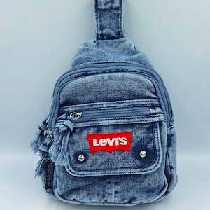 K&ouml;nnte beinhalten: Blaue Jeans-Umh&auml;ngetasche mit rotem Levi's-Logo-Patch. Die Tasche hat ein Rei&szlig;verschluss-Hauptfach, eine Vordertasche und einen kleinen Griff. Der Denim hat ein verwaschenes Aussehen mit ausgefransten Kanten.
