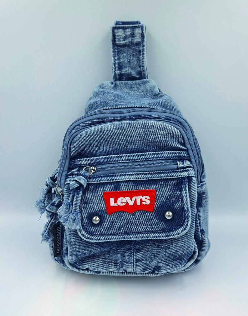 K&ouml;nnte beinhalten: Blaue Jeans-Umh&auml;ngetasche mit rotem Levi's-Logo-Patch. Die Tasche hat ein Rei&szlig;verschluss-Hauptfach, eine Vordertasche und einen kleinen Griff. Der Denim hat ein verwaschenes Aussehen mit ausgefransten Kanten.