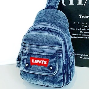 K&ouml;nnte beinhalten: Eine blaue Jeans-Umh&auml;ngetasche mit einer Vordertasche und einem Rei&szlig;verschluss. Die Tasche hat einen roten Levi's-Logoaufn&auml;her. Sie hat einen einzelnen Schultergurt und einen kleinen oberen Griff.