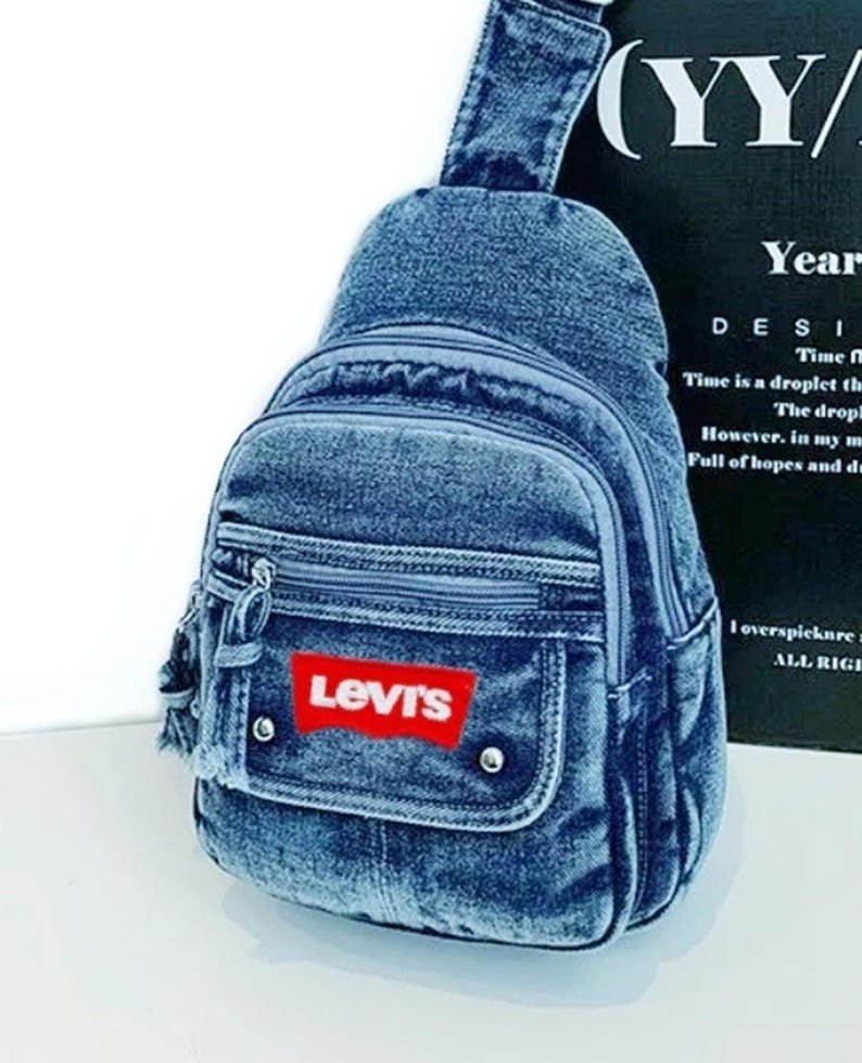 K&ouml;nnte beinhalten: Eine blaue Jeans-Umh&auml;ngetasche mit einer Vordertasche und einem Rei&szlig;verschluss. Die Tasche hat einen roten Levi's-Logoaufn&auml;her. Sie hat einen einzelnen Schultergurt und einen kleinen oberen Griff.