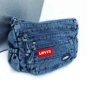 May include: A blue denim crossbody bag featuring a red Levi's brand patch on the front. The bag has multiple zipped compartments with matching blue zip pulls, a side pocket with an elasticated opening, and a silver-toned metal accent.