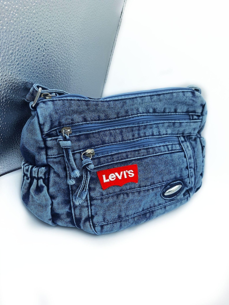 May include: A blue denim crossbody bag featuring a red Levi's brand patch on the front. The bag has multiple zipped compartments with matching blue zip pulls, a side pocket with an elasticated opening, and a silver-toned metal accent.