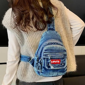 K&ouml;nnte beinhalten: Blaue Jeans-Umh&auml;ngetasche mit einer Vordertasche und einem rot-wei&szlig;en Levi's-Logo. Die Tasche hat ein Hauptfach mit Rei&szlig;verschluss und eine kleinere Rei&szlig;verschlusstasche. Die Tasche wird &uuml;ber der Schulter getragen.