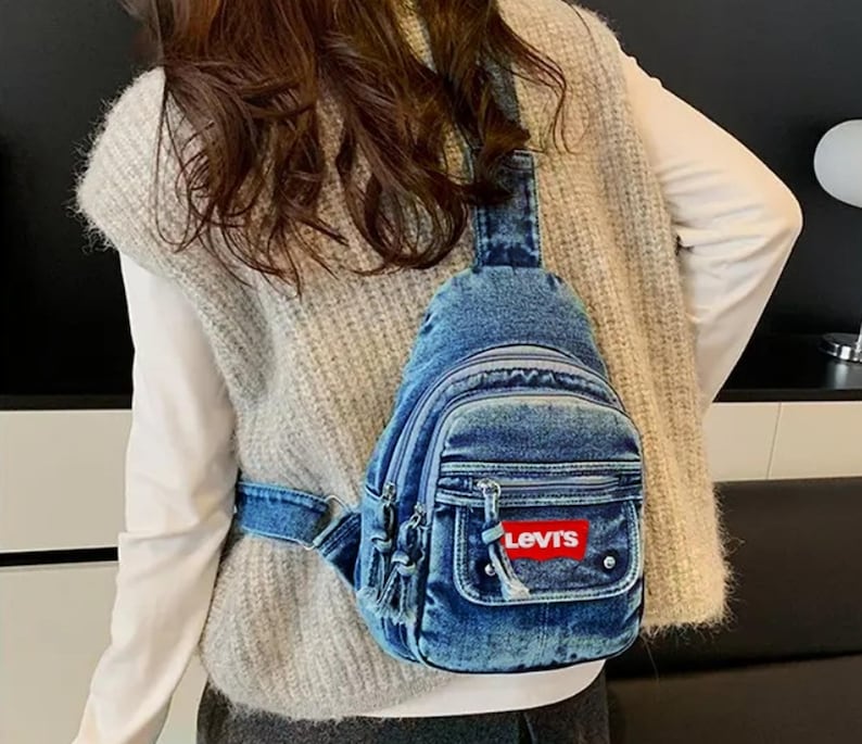 K&ouml;nnte beinhalten: Blaue Jeans-Umh&auml;ngetasche mit einer Vordertasche und einem rot-wei&szlig;en Levi's-Logo. Die Tasche hat ein Hauptfach mit Rei&szlig;verschluss und eine kleinere Rei&szlig;verschlusstasche. Die Tasche wird &uuml;ber der Schulter getragen.