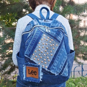 May include: A blue denim backpack featuring a patchwork design with frayed edges, a central panel embellished with metallic studs, and a brown leather patch with the text Lee stitched onto the lower front pocket with silver zippers.