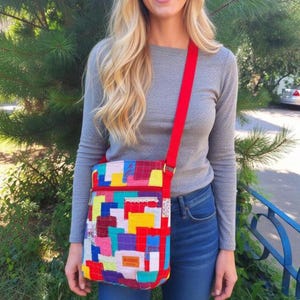 May include: A rectangular crossbody bag featuring a vibrant patchwork design of multicolored fabric squares in red, yellow, blue, and teal. The bag includes a bright red adjustable shoulder strap, a top zipper closure, and a small rectangular label.