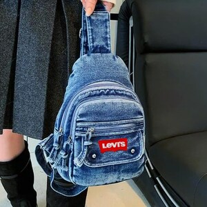 K&ouml;nnte beinhalten: Eine Jeans-Umh&auml;ngetasche mit mehreren Rei&szlig;verschlussf&auml;chern und einer kleinen Vordertasche. Die Tasche hat einen roten Levi's-Logoaufn&auml;her. Die Tasche wird von einer Person getragen, die einen grauen Rock und schwarze Stiefel tr&auml;gt.