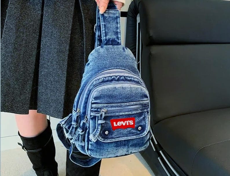 K&ouml;nnte beinhalten: Eine Jeans-Umh&auml;ngetasche mit mehreren Rei&szlig;verschlussf&auml;chern und einer kleinen Vordertasche. Die Tasche hat einen roten Levi's-Logoaufn&auml;her. Die Tasche wird von einer Person getragen, die einen grauen Rock und schwarze Stiefel tr&auml;gt.