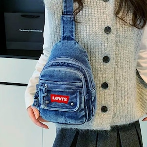 K&ouml;nnte beinhalten: Eine blaue Jeans-Umh&auml;ngetasche mit einer Vordertasche und einem roten Levi's-Logo. Die Tasche hat einen Rei&szlig;verschluss und einen einzelnen Schultergurt. Die Tasche wird &uuml;ber der Schulter getragen.