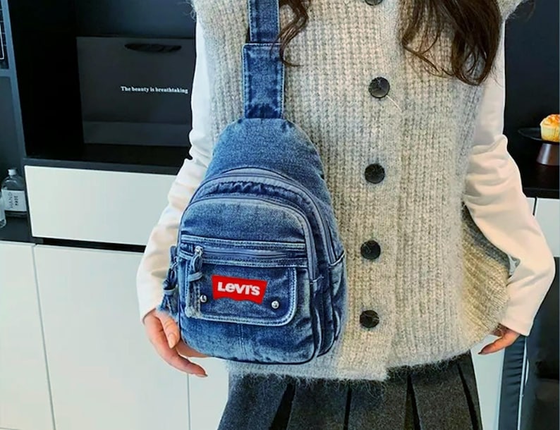 K&ouml;nnte beinhalten: Eine blaue Jeans-Umh&auml;ngetasche mit einer Vordertasche und einem roten Levi's-Logo. Die Tasche hat einen Rei&szlig;verschluss und einen einzelnen Schultergurt. Die Tasche wird &uuml;ber der Schulter getragen.