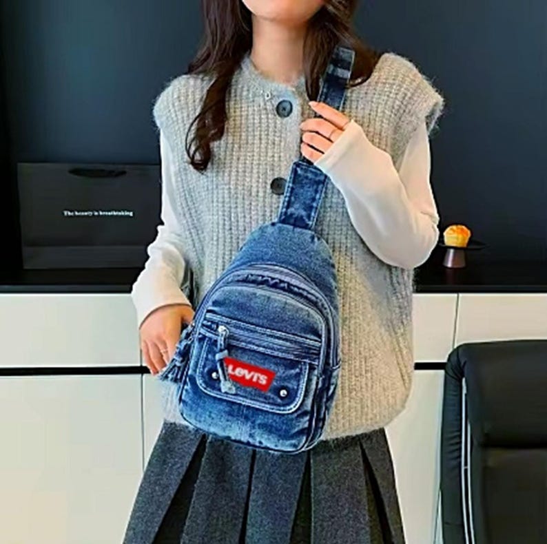 K&ouml;nnte beinhalten: Eine blaue Jeans-Umh&auml;ngetasche mit einer Vordertasche und einem roten "Levis"-Etikett. Die Tasche hat einen einzelnen verstellbaren Riemen und einen Rei&szlig;verschluss. Die Tasche wird &uuml;ber der Schulter getragen.