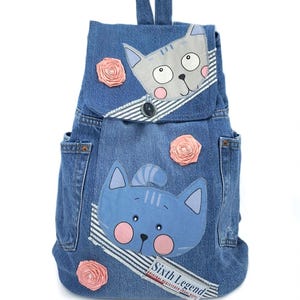 Op de afbeelding: Blauwe denim rugzak met twee kattengezichten, roze accenten en stoffen rozen. De rugzak heeft een klepsluiting met een knoop en twee zijzakken. De tekst "Sixth Legend" is zichtbaar op de voorkant.