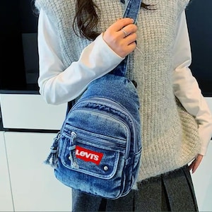 K&ouml;nnte beinhalten: Eine blaue Jeans-Umh&auml;ngetasche mit mehreren Rei&szlig;verschlussf&auml;chern und einer kleinen Vordertasche. Die Tasche hat das Levi's-Logo in einem roten Rechteck. Sie hat einen Schultergurt und wird quer &uuml;ber dem K&ouml;rper getragen.