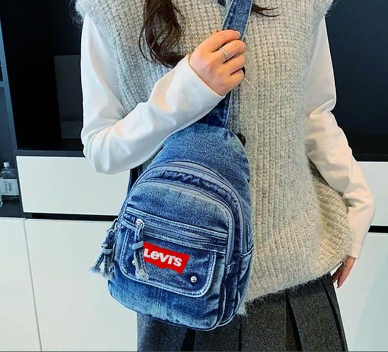 K&ouml;nnte beinhalten: Eine blaue Jeans-Umh&auml;ngetasche mit mehreren Rei&szlig;verschlussf&auml;chern und einer kleinen Vordertasche. Die Tasche hat das Levi's-Logo in einem roten Rechteck. Sie hat einen Schultergurt und wird quer &uuml;ber dem K&ouml;rper getragen.
