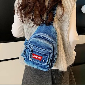 K&ouml;nnte beinhalten: Eine blaue Jeans-Umh&auml;ngetasche mit einem rot-wei&szlig;en Logo-Patch. Die Tasche hat ein Hauptfach mit Rei&szlig;verschluss, eine Vordertasche und eine kleine Seitentasche. Die Tasche wird &uuml;ber der Schulter getragen.