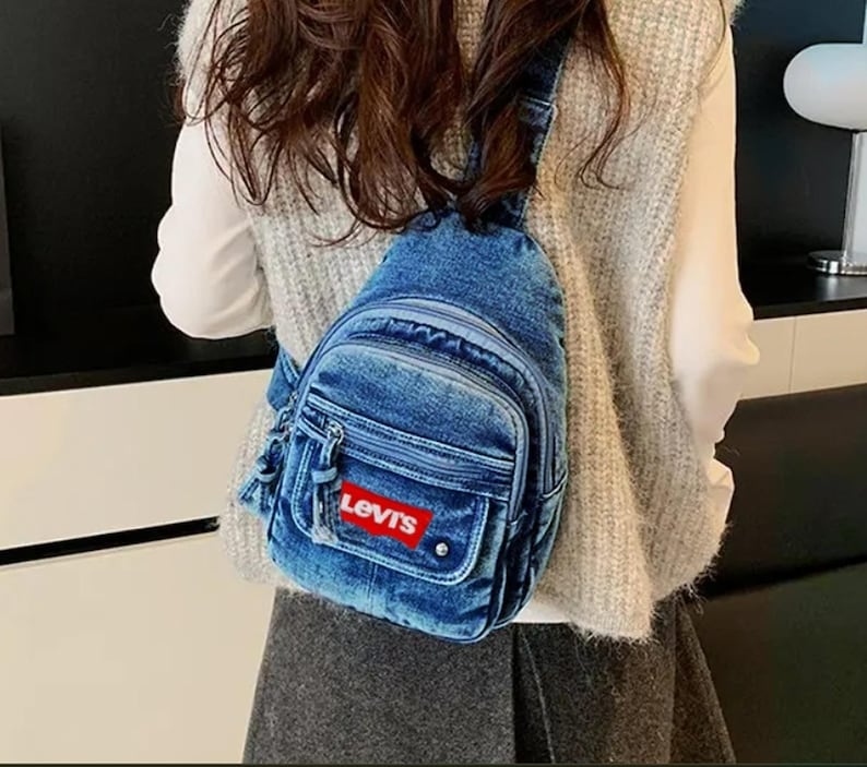 K&ouml;nnte beinhalten: Eine blaue Jeans-Umh&auml;ngetasche mit einem rot-wei&szlig;en Logo-Patch. Die Tasche hat ein Hauptfach mit Rei&szlig;verschluss, eine Vordertasche und eine kleine Seitentasche. Die Tasche wird &uuml;ber der Schulter getragen.