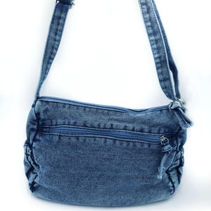 May include: A medium blue denim crossbody bag featuring a rectangular main compartment with a top zip closure, a horizontal front zipped pocket, and gathered side panels. The bag includes an adjustable denim shoulder strap with metal hardware.