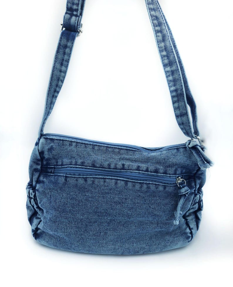 May include: A medium blue denim crossbody bag featuring a rectangular main compartment with a top zip closure, a horizontal front zipped pocket, and gathered side panels. The bag includes an adjustable denim shoulder strap with metal hardware.