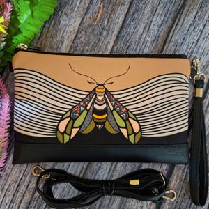 May include: A black and tan clutch with a colorful moth design. The clutch features a black wrist strap and a detachable black shoulder strap. The moth has green, orange, and yellow accents. The background has a wavy pattern.