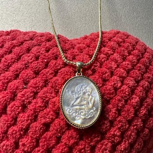 Op de afbeelding: Een goudkleurige ketting met een ovale hanger met een gebeeldhouwd engelontwerp. De hanger heeft een parelmoer-achtige uitstraling en is gezet in een goudkleurig montuur. De ketting ligt op een rode, getextureerde ondergrond.