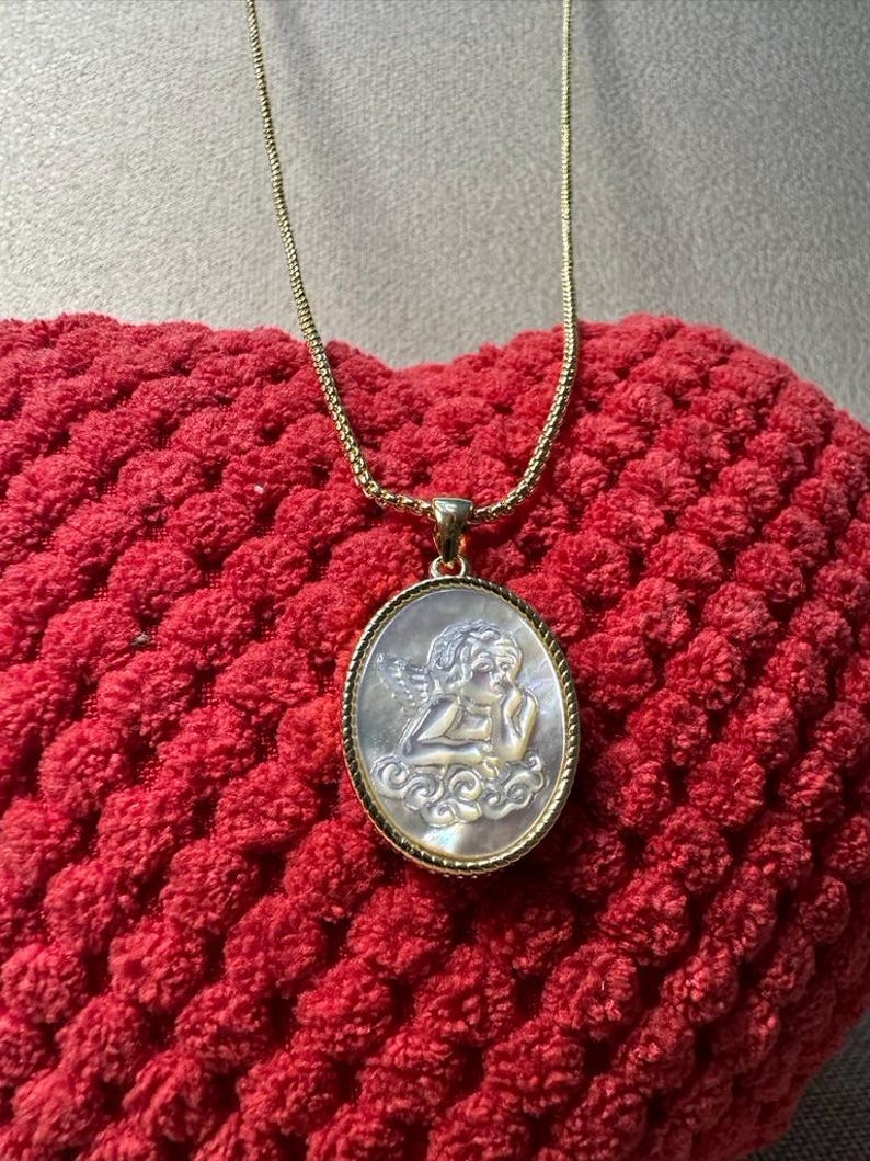 Op de afbeelding: Een goudkleurige ketting met een ovale hanger met een gebeeldhouwd engelontwerp. De hanger heeft een parelmoer-achtige uitstraling en is gezet in een goudkleurig montuur. De ketting ligt op een rode, getextureerde ondergrond.