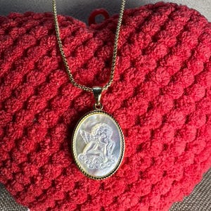 Op de afbeelding: Een goudkleurige ketting met een ovale hanger met een gebeeldhouwd engelontwerp. De hanger heeft een parelmoeren achtergrond en een goudkleurige lijst. De ketting wordt gepresenteerd op een rode, getextureerde ondergrond.