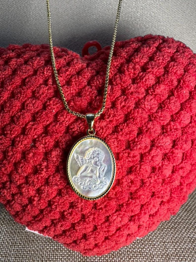 Op de afbeelding: Een goudkleurige ketting met een ovale hanger met een gebeeldhouwd engelontwerp. De hanger heeft een parelmoeren achtergrond en een goudkleurige lijst. De ketting wordt gepresenteerd op een rode, getextureerde ondergrond.
