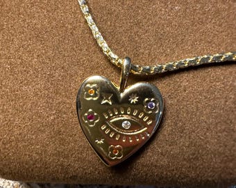 May include: Gold heart-shaped pendant necklace with a detailed eye design and colorful gemstones. The necklace is on a brown velvet surface.