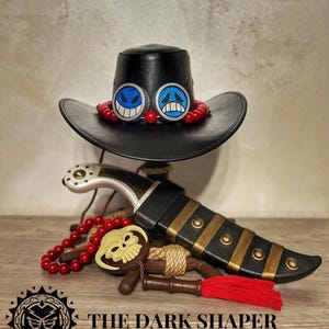 Puede incluir: Sombrero de cuero negro con emblemas faciales azules y blancos y cuentas rojas. Un cuchillo decorativo con una vaina negra y detalles dorados, un collar de cuentas rojas y un dije en forma de calavera. El texto "THE DARK SHAPER" está en la parte inferior.