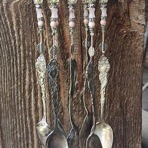 May include: A wind chime featuring five silver-toned vintage spoons hanging from a circular base. Each spoon is suspended by a vertical strand of light pink beads, clear glass beads with floral patterns, and intricate silver-toned metal spacers.