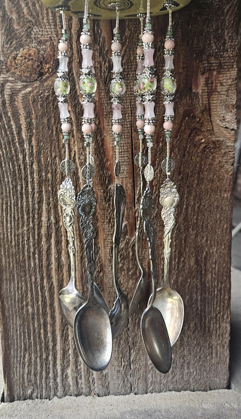 May include: A wind chime featuring five silver-toned vintage spoons hanging from a circular base. Each spoon is suspended by a vertical strand of light pink beads, clear glass beads with floral patterns, and intricate silver-toned metal spacers.