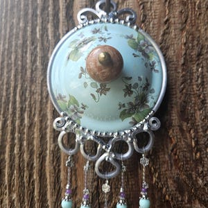 May include: A decorative wall hanging featuring a light blue circular ceramic plate with a floral pattern and a central brown knob. The plate is framed in silver metal with ornate scrollwork and five dangling strands of beads in light blue and purple.