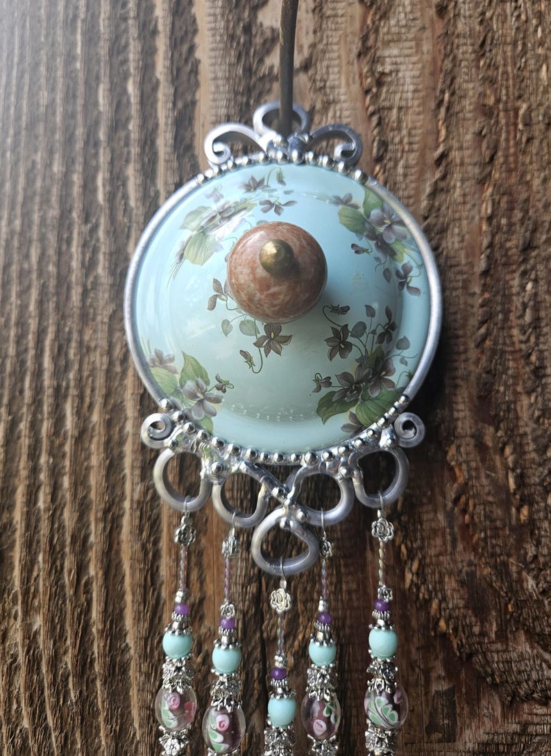 May include: A decorative wall hanging featuring a light blue circular ceramic plate with a floral pattern and a central brown knob. The plate is framed in silver metal with ornate scrollwork and five dangling strands of beads in light blue and purple.