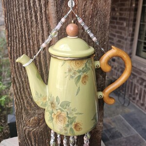 May include: A pale yellow enamel teapot featuring a floral rose pattern in shades of yellow and green. The teapot has a wooden handle and a wooden knob on the lid. It is suspended by a decorative wire hanger adorned with pink and silver beads.
