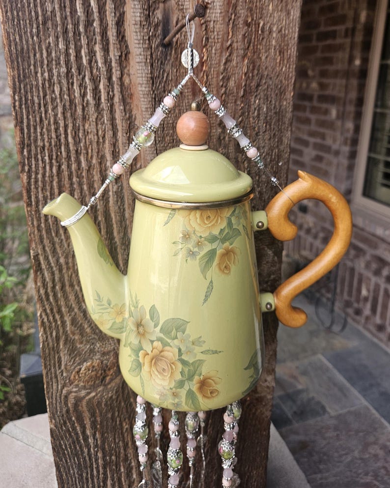 May include: A pale yellow enamel teapot featuring a floral rose pattern in shades of yellow and green. The teapot has a wooden handle and a wooden knob on the lid. It is suspended by a decorative wire hanger adorned with pink and silver beads.