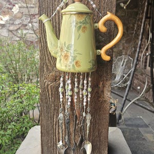 May include: A decorative wind chime featuring a light green teapot with a yellow floral pattern, a wooden handle, and a lid. Strands of pink and green beads hang from the base, interspersed with several silver-toned metal spoons.