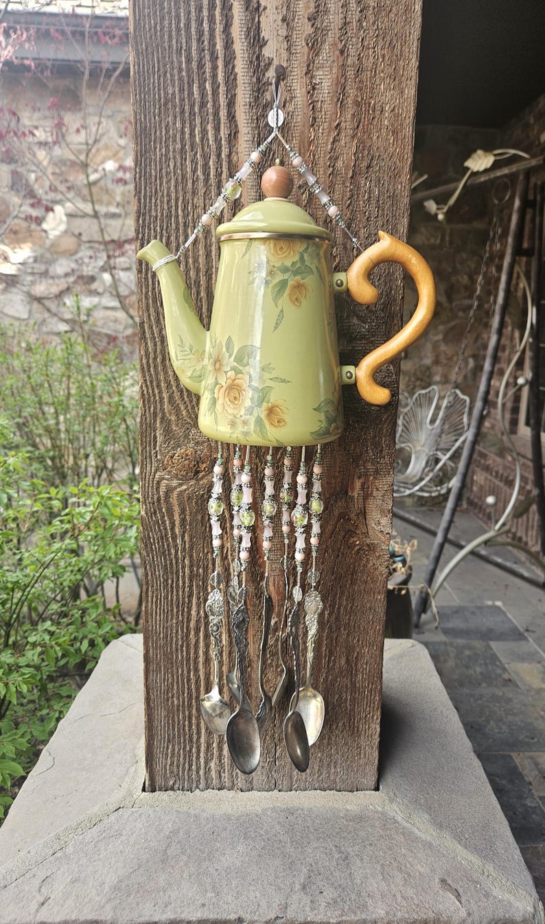 May include: A decorative wind chime featuring a light green teapot with a yellow floral pattern, a wooden handle, and a lid. Strands of pink and green beads hang from the base, interspersed with several silver-toned metal spoons.