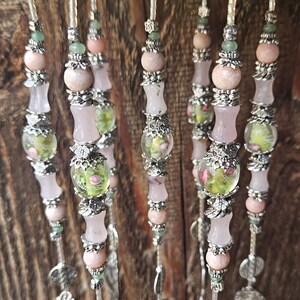 May include: A series of vertical beaded strands featuring pale pink and translucent beads, interspersed with silver-toned metal floral spacers and clear glass beads containing green and pink floral patterns, ending with silver-toned metal charms.