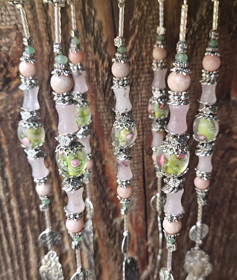 May include: A series of vertical beaded strands featuring pale pink and translucent beads, interspersed with silver-toned metal floral spacers and clear glass beads containing green and pink floral patterns, ending with silver-toned metal charms.