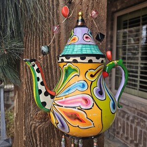 May include: A colorful, hand-painted teapot wind chime. The teapot is yellow with vibrant floral designs in green, blue, purple, and orange. It has a green handle and spout, and is adorned with hanging beaded strands and decorative beads.