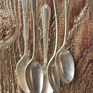 May include: A wind chime constructed from five vintage silver-toned metal spoons hanging vertically. Each spoon features an ornate embossed handle design and is suspended by a thin string adorned with small silver-colored metal beads and accents.