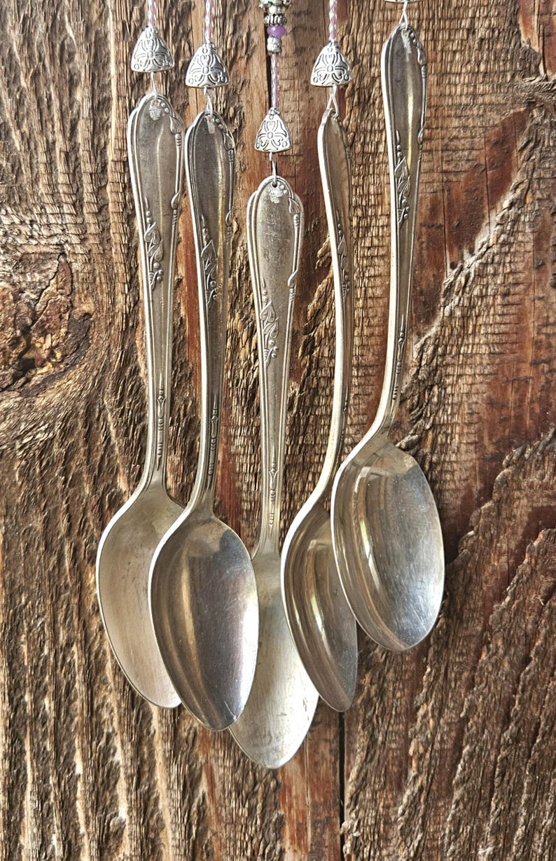 May include: A wind chime constructed from five vintage silver-toned metal spoons hanging vertically. Each spoon features an ornate embossed handle design and is suspended by a thin string adorned with small silver-colored metal beads and accents.