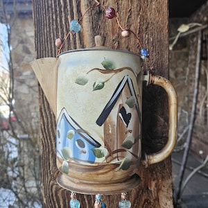 May include: A decorative metal teapot wind chime featuring hand-painted illustrations of two birdhouses, one blue and one brown, surrounded by green leaves. The teapot is suspended by a copper wire adorned with colorful glass beads and charms.
