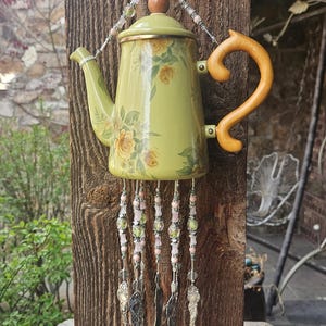 May include: A decorative wind chime crafted from a light green teapot featuring yellow floral patterns, a wooden handle, and a lid. Five silver-toned metal spoons hang from the base of the teapot, suspended by strings of pink and green beads.